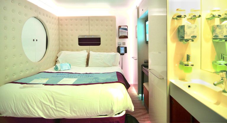 Cabine Studio pour une personne à bord du Norwegian Epic : lit double contre une paroi capitonnée blanche, hublot décoratif rond, lavabo et miroir au premier plan