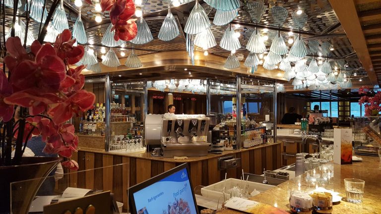 Le Palatino Grand Bar du Costa Favolosa : comptoir en bois, machine à expresso, bouteilles d'alcool et lustres en verre en forme de coquillage
