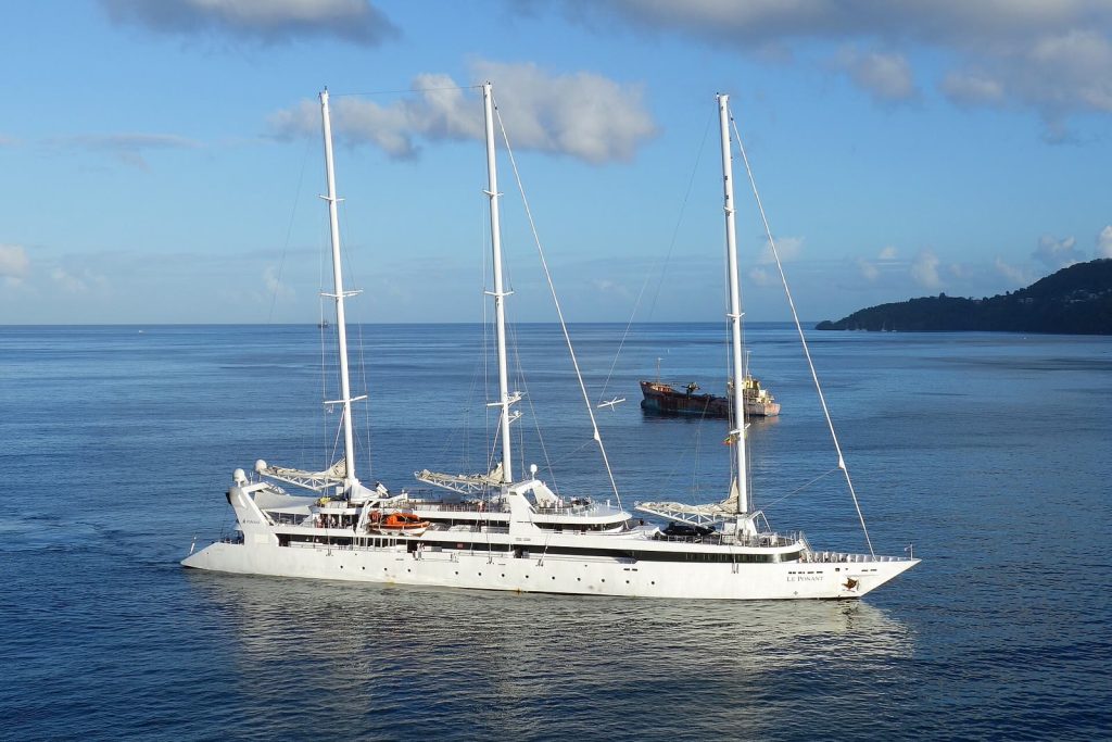 Le voilier trois-mâts Le Ponant, coque blanche et voiles ferlées, naviguant de profil sur une mer calme près d'une côte boisée