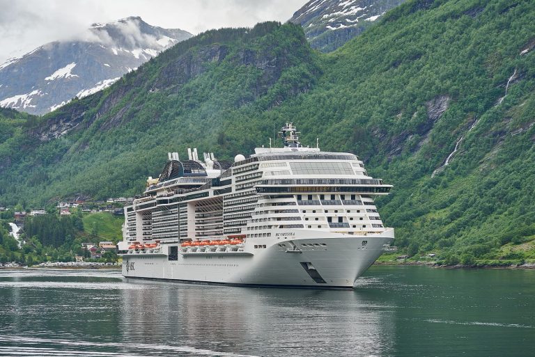 Le paquebot MSC Virtuosa vu de trois quarts avant dans le Geirangerfjord, entre des montagnes boisées aux sommets enneigés et une cascade
