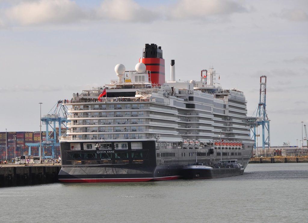 Le paquebot Queen Anne de Cunard, coque noire à liseré rouge et cheminée rouge et noire, amarré à un quai du port de Zeebrugge, vu par l'arrière avec son nom sur la poupe