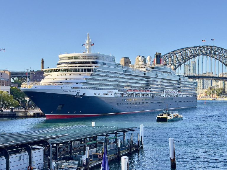 Le paquebot Queen Elizabeth de Cunard, coque bleu marine et liseré rouge, amarré dans le port de Sydney avec le Harbour Bridge en arrière-plan