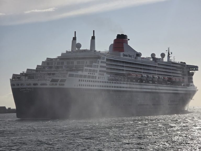 Le paquebot Queen Mary 2 de Cunard, coque noire et cheminée rouge, quittant le port du Havre dans un nuage d'embruns projetés par un remorqueur