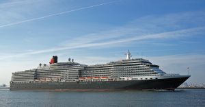 Le paquebot Queen Victoria de Cunard naviguant de profil, coque bleu nuit, superstructure blanche et cheminée rouge et noire, sous un ciel bleu
