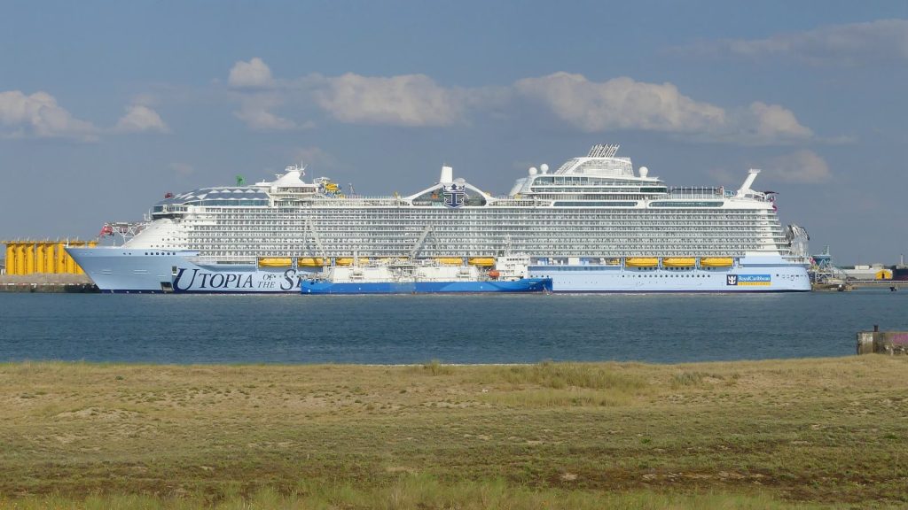 Le paquebot Utopia of the Seas de Royal Caribbean à quai à La Rochelle-La Pallice, vu de profil depuis la côte, avec un petit navire bleu le long de sa coque